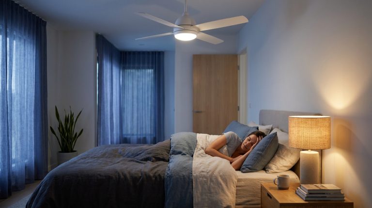 Chambre la nuit : femme endormie sous ventilateur de plafond blanc, lumière chaude d'une lampe de chevet, porte fermée visible.