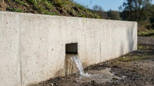 Mur de soutènement en béton avec une barbacane carrée d'où s'écoule de l'eau sur un sol terreux. L'herbe pousse sur la butte.