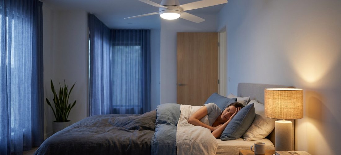 Chambre la nuit : femme endormie sous ventilateur de plafond blanc, lumière chaude d'une lampe de chevet, porte fermée visible.