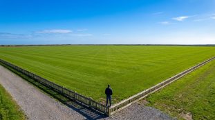 Vue aérienne d'un homme sur une clôture, face à un immense champ de gazon vert parfaitement tondu sous un ciel bleu.