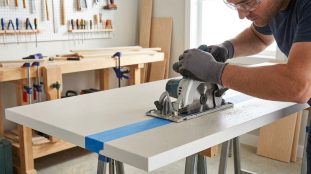Homme coupant un plan de travail clair avec une scie circulaire. Il porte gants et lunettes. Du ruban adhésif bleu marque la ligne de coupe.