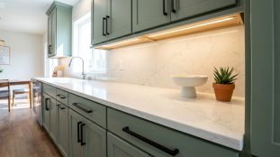 Une cuisine moderne avec des armoires vert sauge, des poignées noires, des comptoirs en quartz blanc et un éclairage sous armoire.