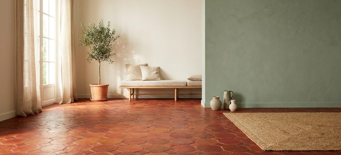 Pièce lumineuse avec tomettes rouges hexagonales. Murs vert sauge et crème, canapé en bois, olivier, tapis et vases en terre.