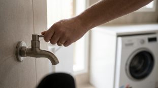 Gros plan sur une main manipulant un robinet mural en métal pour machine, avec des gouttes d'eau. Machine à laver floue en arrière-plan.