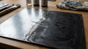 Plaque de zinc en cours de patination, montrant une transition d'un aspect brillant à un effet vieilli sombre. Bouteilles et pinceaux en arrière-plan.