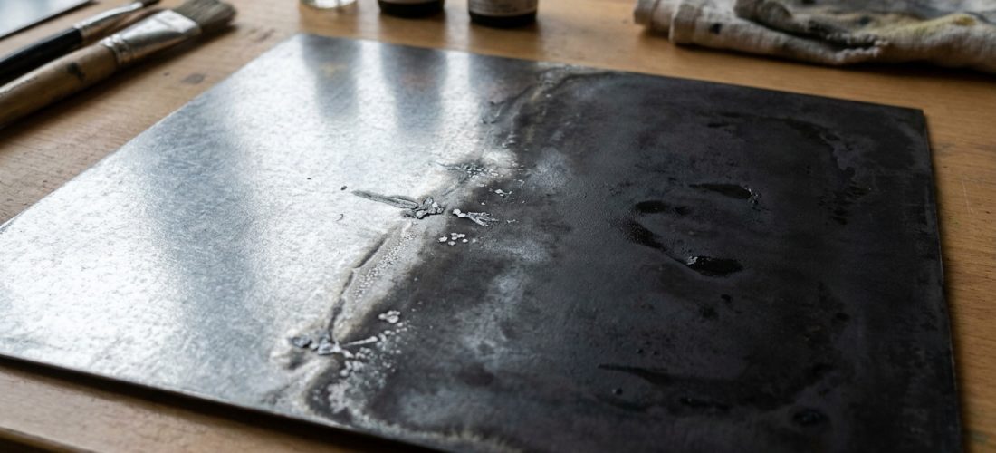 Plaque de zinc en cours de patination, montrant une transition d'un aspect brillant à un effet vieilli sombre. Bouteilles et pinceaux en arrière-plan.