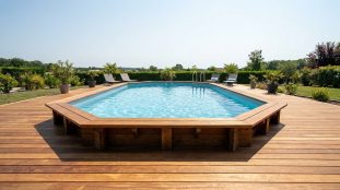Piscine octogonale hors-sol avec liner bleu, intégrée à une large terrasse en bois clair. Transats, plantes et verdure sous un ciel ensoleillé.