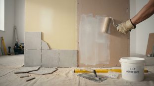 Vue rapprochée d'un mur en rénovation. Une partie est jaune avec des carreaux gris posés; une main peint l'autre partie en blanc. Outils au sol.