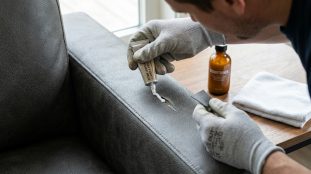 Personne en gants appliquant une pâte de réparation sur une déchirure d'un accoudoir de canapé gris en skaï ou vinyle.